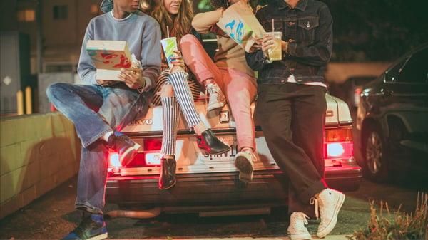 Three young people leaning against the trunk of the car while eating Taco Bell.