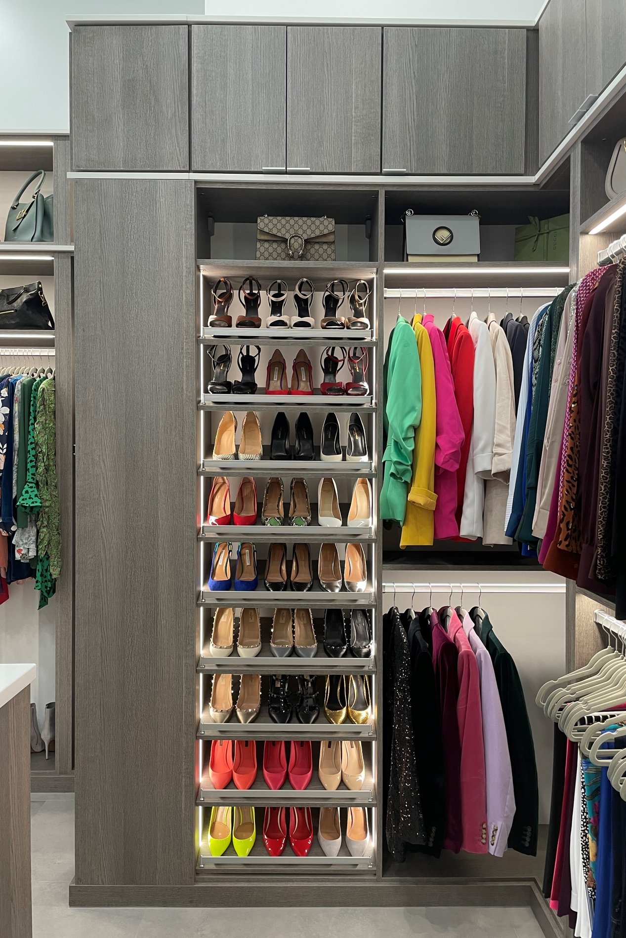 A walk-in closet in ash finish featuring vertical and horizontal lighting, angled shoe storage, and doors with finger pulls.