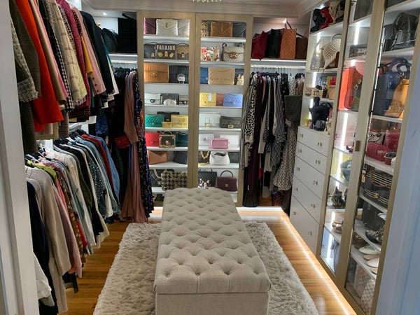 Walk-in closet in white with hanging clothes and purses behind glass doors with center ottoman
