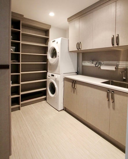 Custom laundry room with stacked washer/dryer, sink and light grey wood cabinets and shelves.