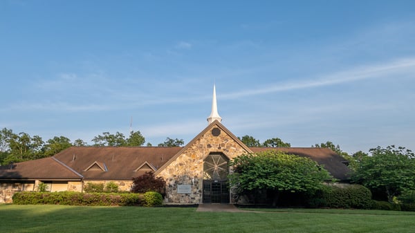 The Church of Jesus Christ of Latter-day Saints meetinghouse in Signal Mountain, Tennessee. The viewer is presented with a stucco and stone building with pitched roof, large round window over the front glass door, and a white spire. The Church of Jesus Christ of Latter-day Saints is carved into granite and mounted to the left of the front glass door. The meeting house sits directly across from the Signal Mountain Post Office.