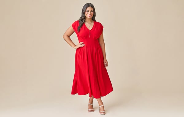 Woman is wearing a plus size red midi dress, and is posing with one hand on her hip in front of a tan background.