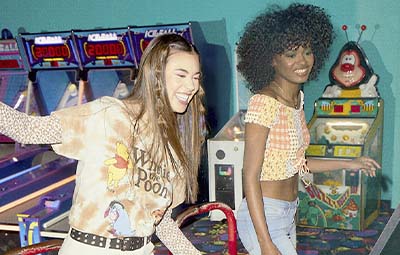 Two female models; one wearing a crop top, the other wearing a long sleeve graphic t-shirt, playing in an arcade.