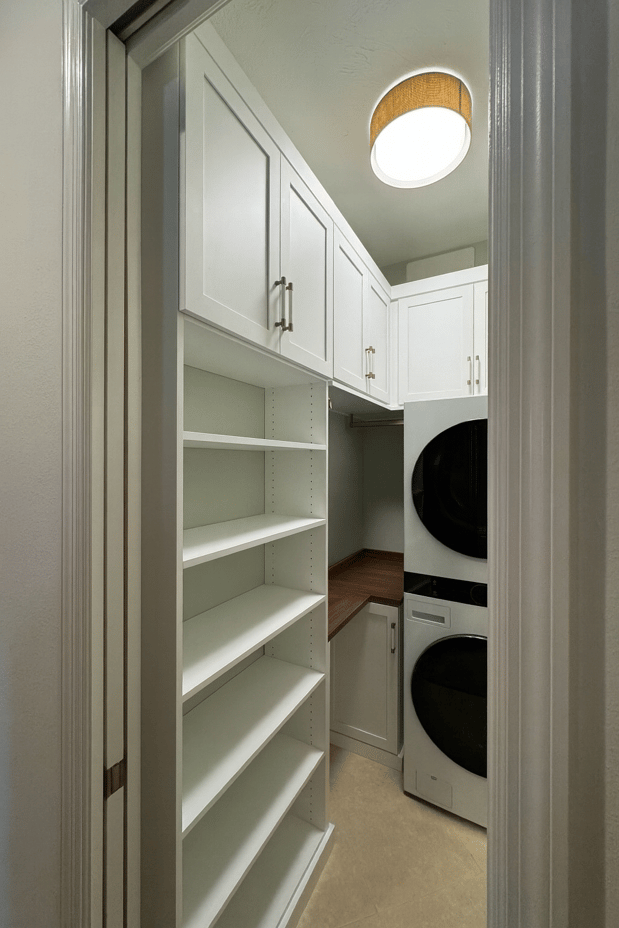 Compact laundry room with custom white shelving, floor-to-ceiling storage, cabinetry, and stacked washer and dryer.