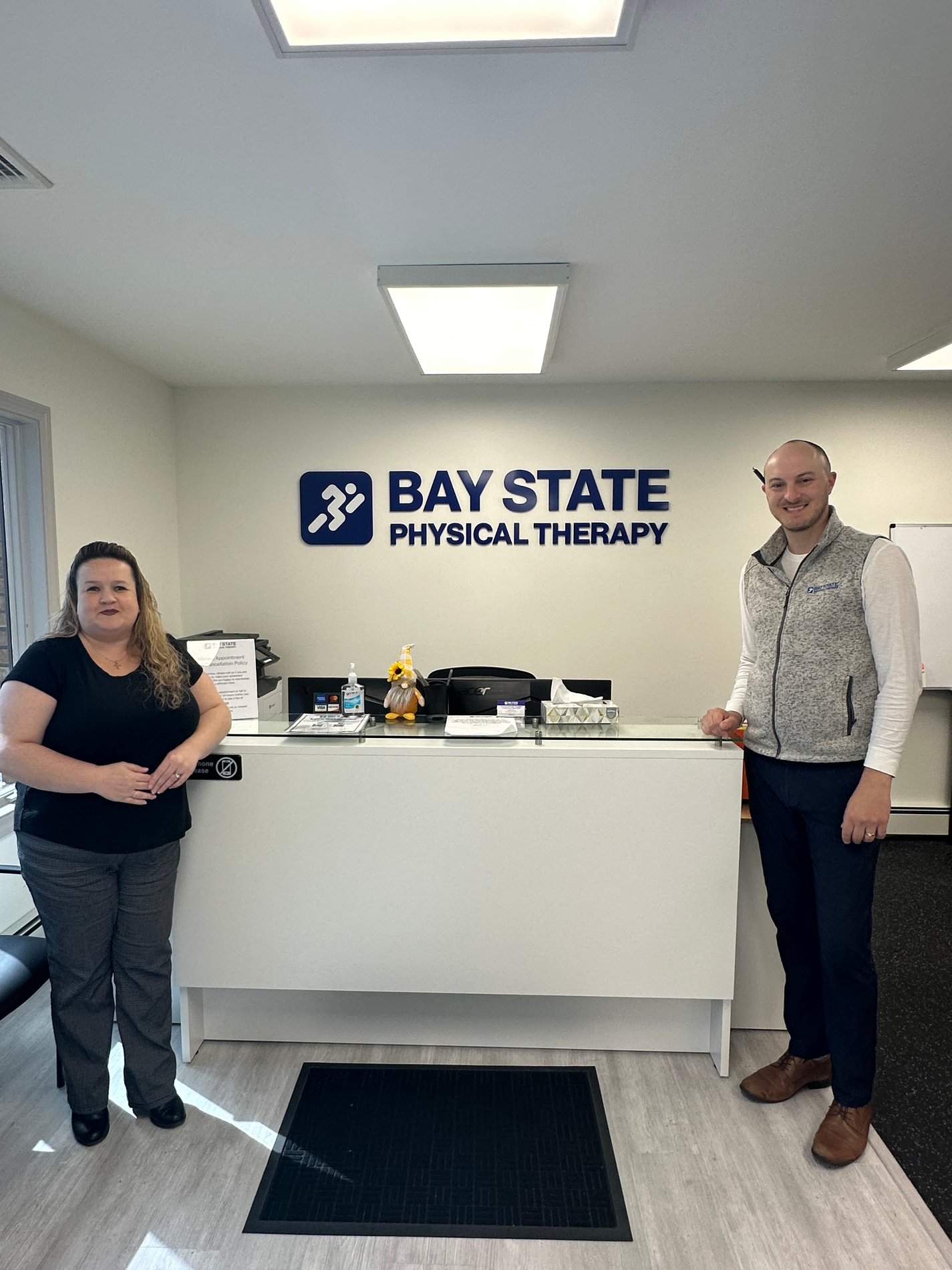 RI-WestWarwick-BSPT front desk and waiting room area Bay State PT logo on wall above desk with staff members around.