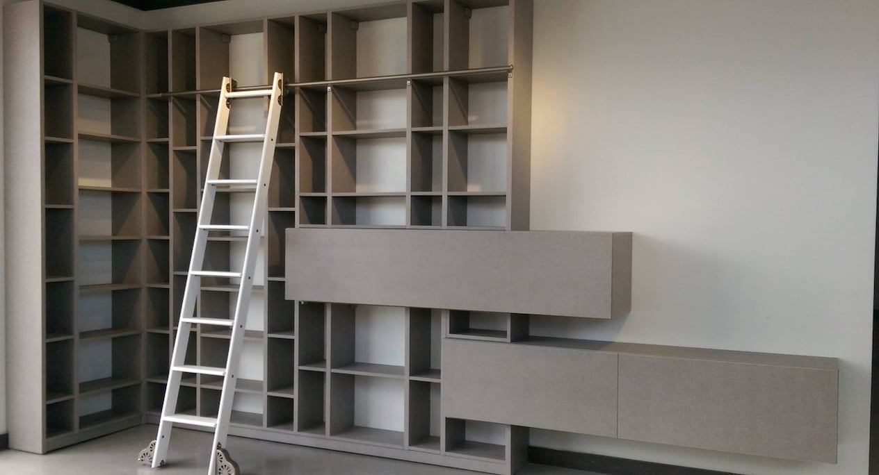A modern built-in wall shelving unit with multiple square cubbies in a gray finish. A white rolling ladder leans against the shelves