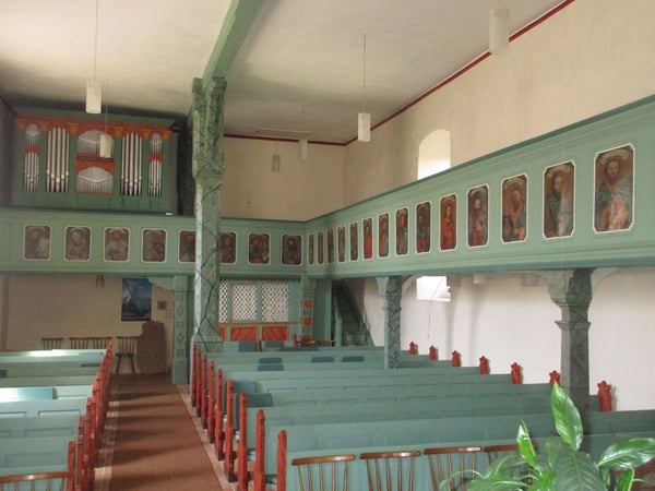 Das Bild zeigt das Innere der Evangelische Kirche Kirtorf-Gleimenhain. Die prägenden farben sind beige und türkis. Außerdem sind eine Gemälde an der Wand zu erkennen.