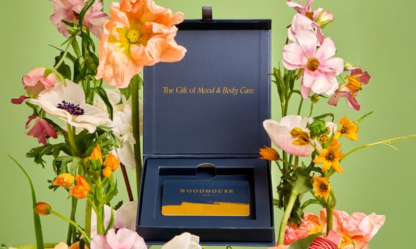 Woodhouse Spa Gift Card Box open to reveal gift card in front of green background and flowers on the sides
