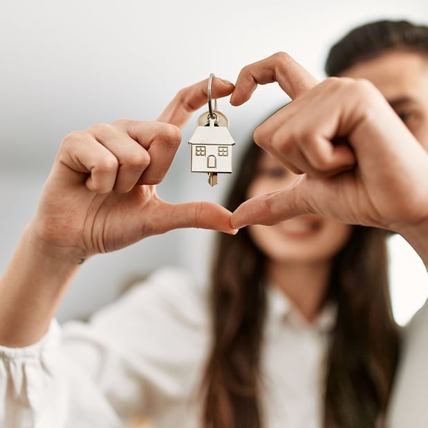 A woman and man making a heart shape with their fingers and a house key ring is hanging from her index finger.