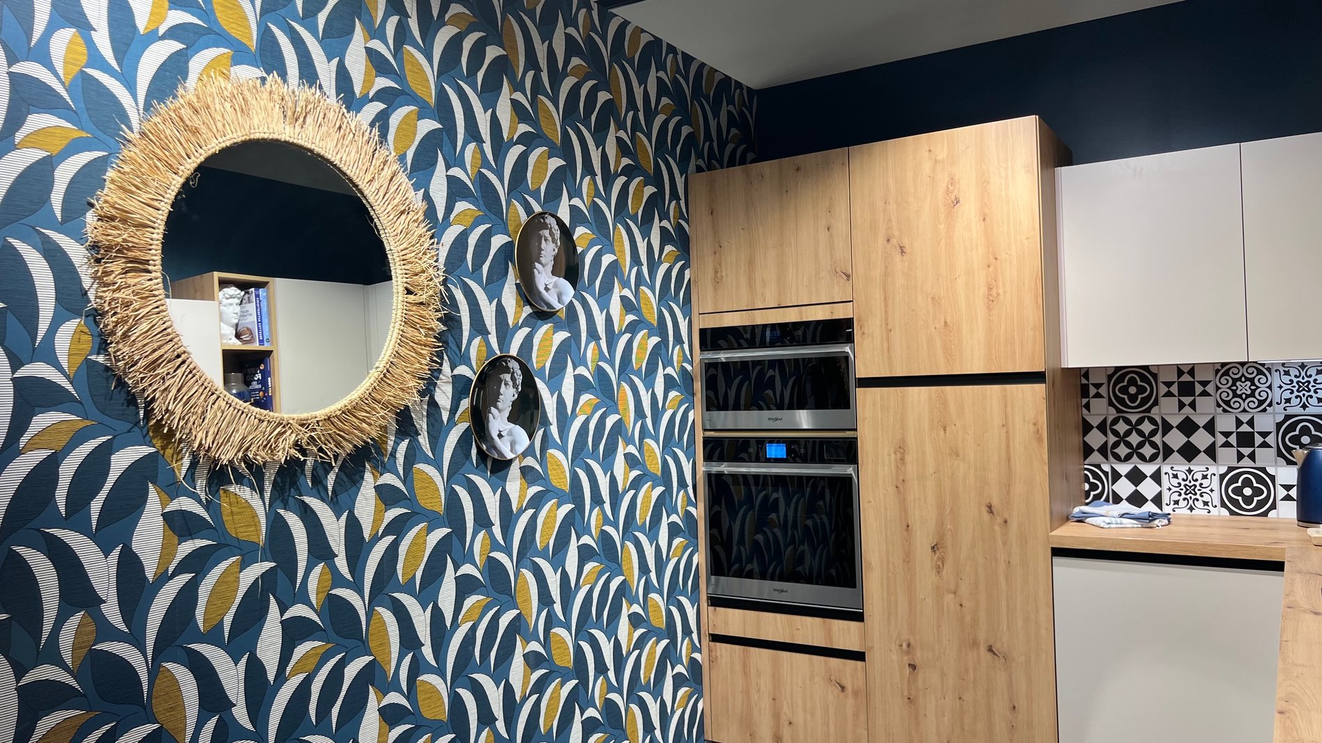 Côté gauche de la cuisine. Mur en motif bleu, jaune et banc avec un petit miroir en forme ovale.
Deux meuble en bois accompagné d'un four connecté.