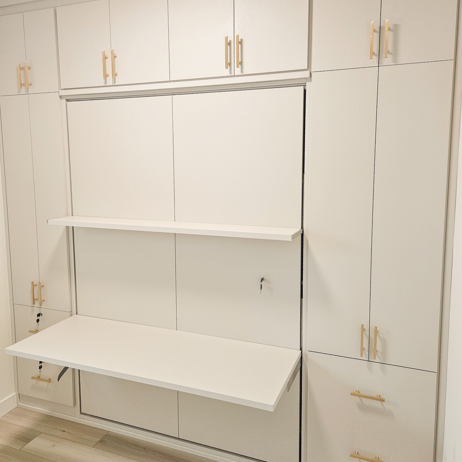 Custom wall bed in a white finish with gold hardware. The desk and shelf on the front fold down underneath the bed when the bed is in use.