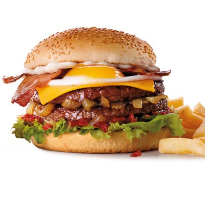 Beef burger with two rashers of bacon, an egg, cheese, mayo, onions, gherkins, mixed lettuce and relish in front of chips against white background.