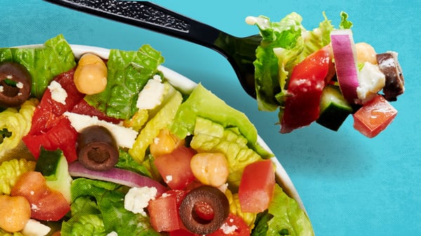 A Salad from MOD Pizza with olives and tomatoes is on a blue background, a plastic fork is holding a bite of the salad.