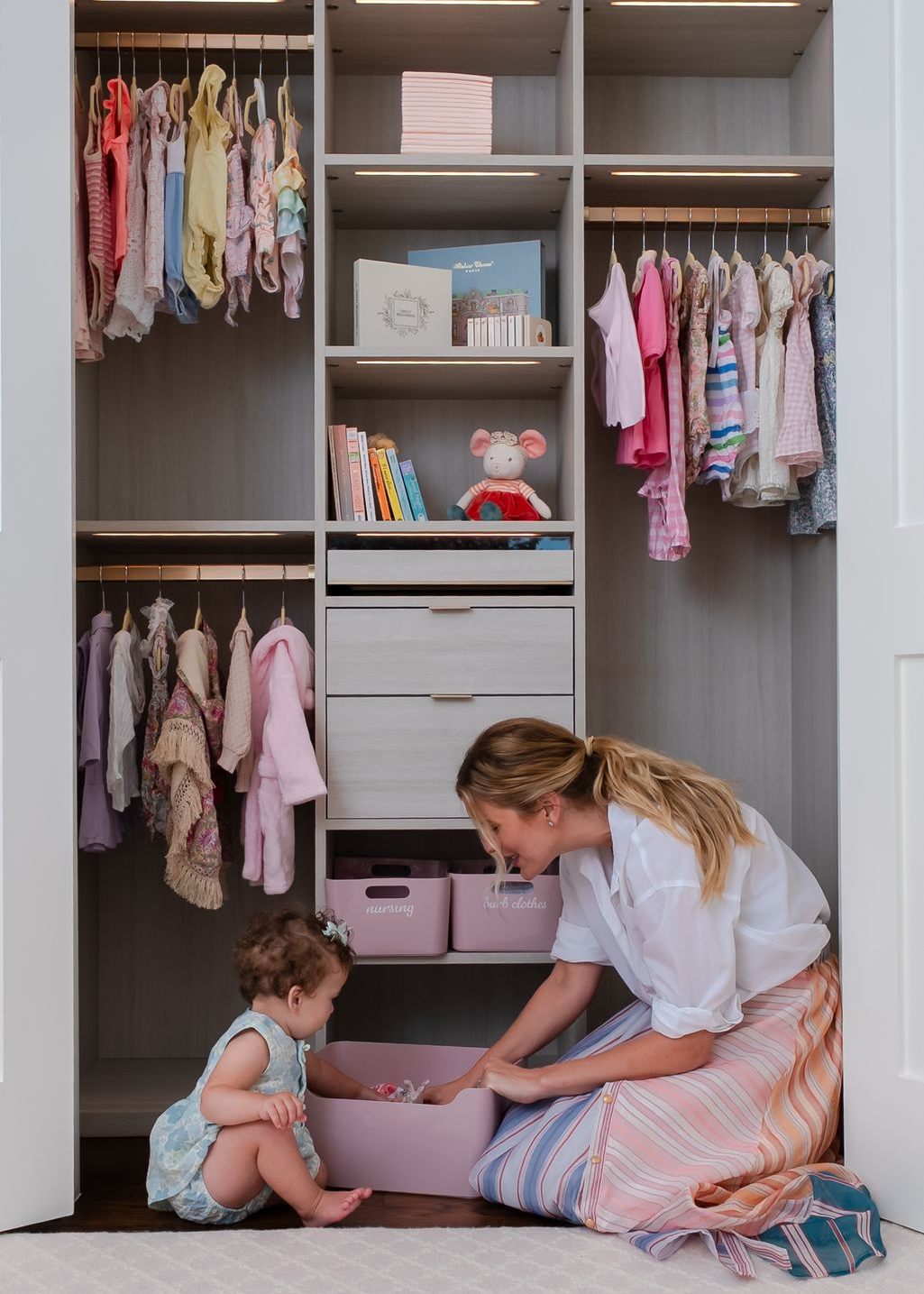 woman kneels beside a toddler on the floor of a nursery closet