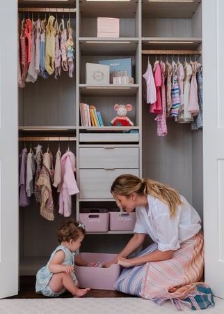 woman kneels beside a toddler on the floor of a nursery closet
