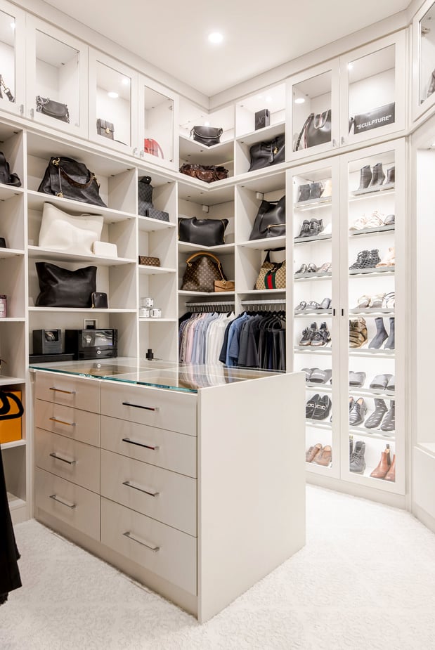 Walk-in Closet designed in a all white finish with floor to ceiling storage, open shelving, shoe shelves behind glass doors, and a center island. Lighting adds an elevated touch to the whole system.
