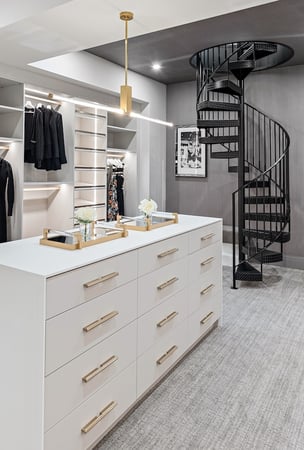 Custom walk-in closet by California Closets Greater Detroit in a home in Romeo, Michigan. featuring white built-in cabinetry with hanging rods, shelving, drawers, and glass-front upper cabinets, along with a contrasting dark center island with drawers.