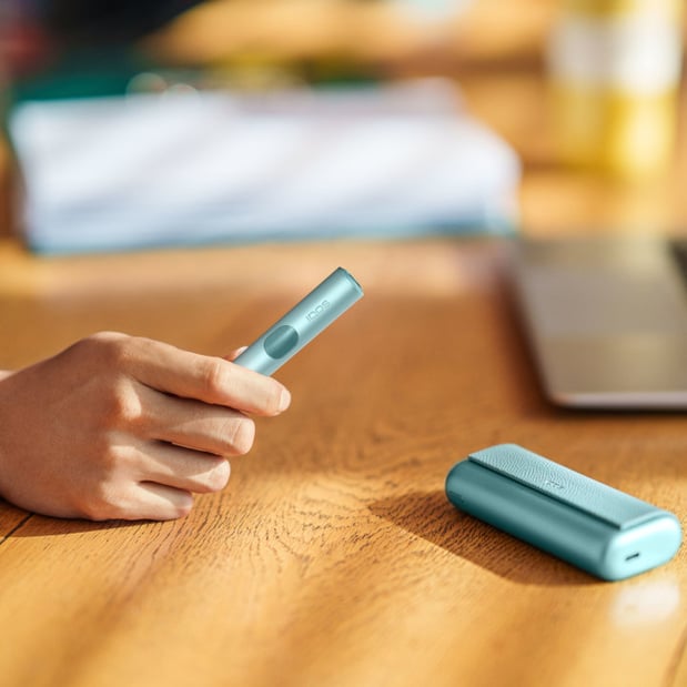 Osoba trzyma urządzenie IQOS Iluma, podczas gdy jego ładowarka leży na drewnianym stole obok laptopa.