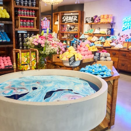 Inside a LUSH North America store where piles of bath bombs sit on a wooden table, organized by color. Against the wall are colorful products and in the foreground is a large stone tub swirling with colorful, foaming water.