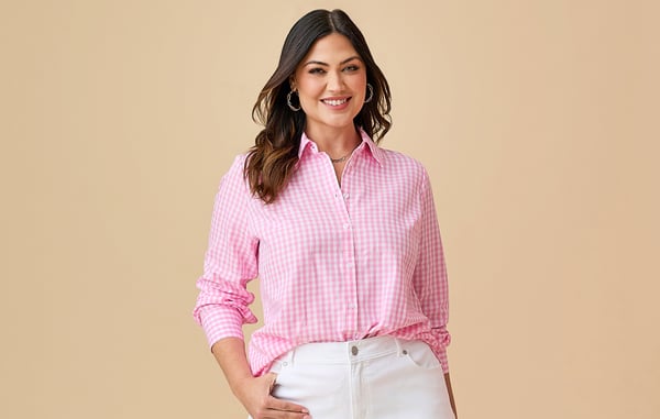 Woman is wearing a plus size pink gingham button down shirt and white jeans, and is posing with one hand in her pocket in front of a tan background.