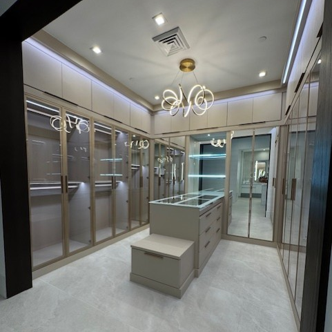 Primary closet in Stone with matching backing, featuring suspended poles, gold accessories, gold aluminum framed mirrored doors with concealed shoe shelving, Elusion framed countertop, integrated lighting, and a secure drawer safe.