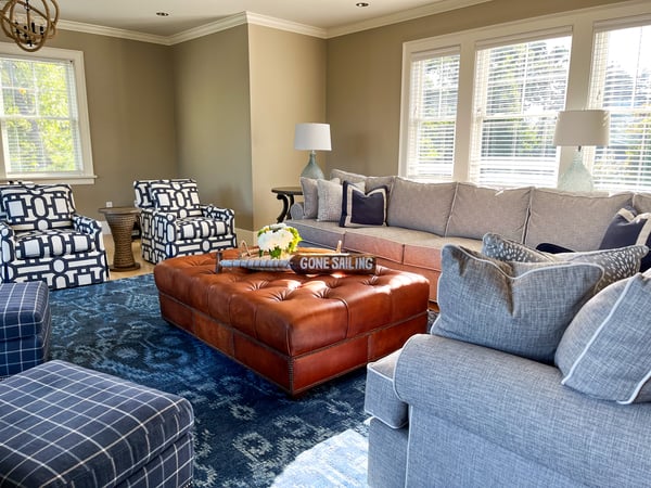 A comfortable living room with grey couch, blue and white geometric print armchairs, a blue patterned rug, and a leather ottoman by designer Melonie Montiero for Cabot House Furniture.