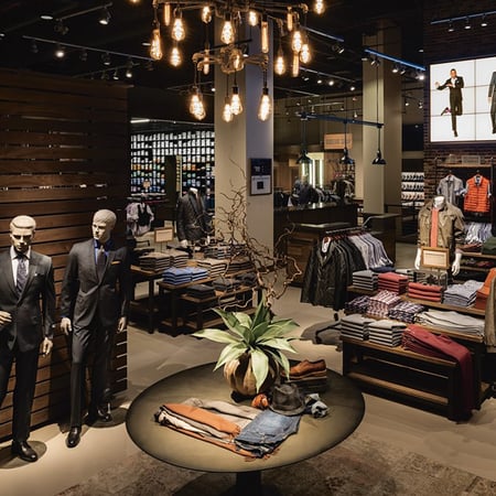 Inside a Tailored Brands Location, two mannequins stand in dark gray suits, and jeans and jacket outfit sits on a table in the foreground and lines of neatly folded button down shirts line a table.
