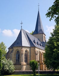 Zu sehen ist die Kirche Ober-Hilbersheim. Eine Turmspitze ragt in den blauen Himmel und in der Umgebung wachsen Bäume.