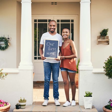 A man and a woman standing in front of a house. The man is holding a chalkboard that reads 