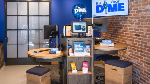 Inside a Dime community bank location, a tv shows "It's Your Dime" on the screen, there's a Dime branded balloon and various pamphlets dividing two computer stations with cushioned benches for Dime Employees and customers