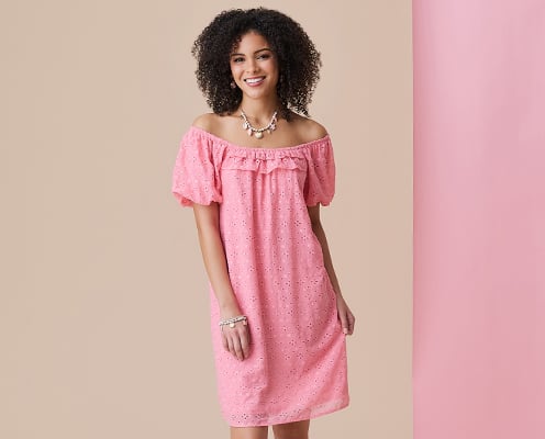 Woman is wearing a pink off the shoulder spring dress and posing in front of a tan and pink background.