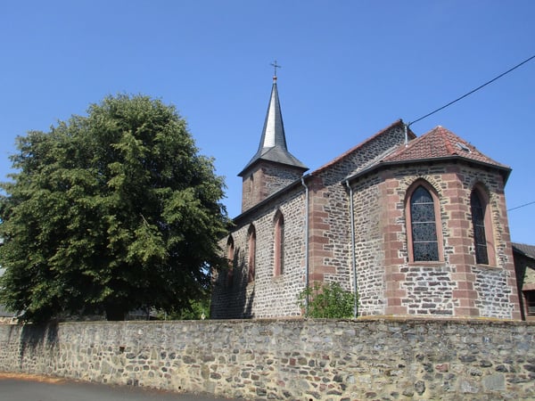 Das Bild zeigt die Evangelische Kirche Arnshain von außen mit altem Gemäuer, einem spitzen Kirchturm. Links der Kirche steht ein großer grüner Baum.